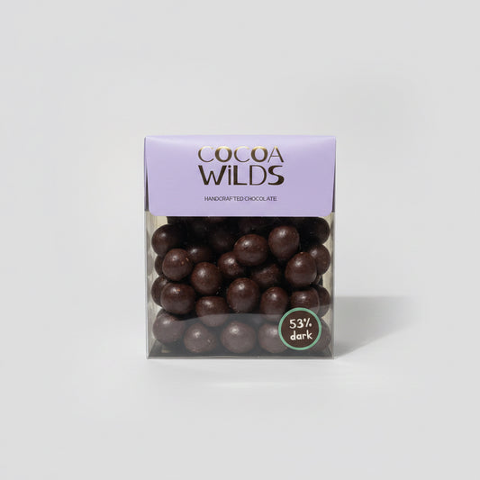 A box of Cocoa Wilds Handcrafted Chocolate on a white countertop. The packaging features a lavender purple top flap with the brand name 'COCOA WILDS' in brown lettering, and a clear lower section showing dark chocolate-covered macadamias inside.